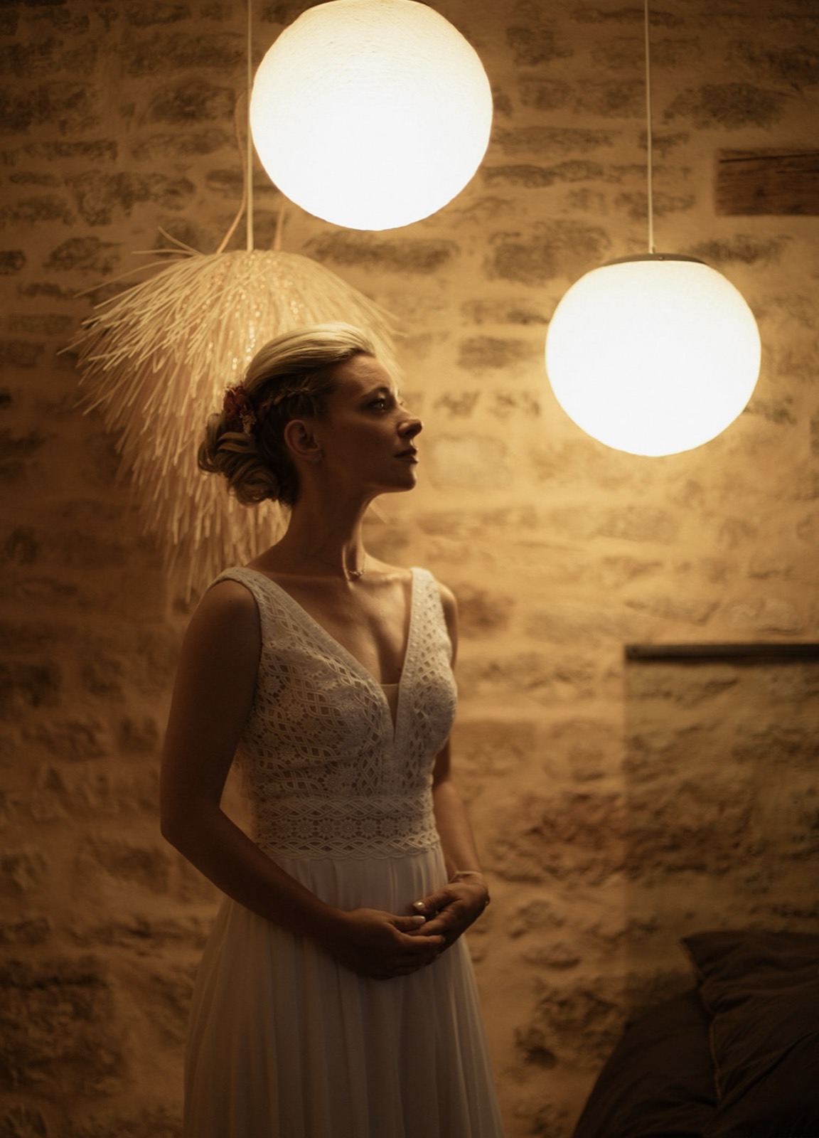 Une mariée coiffée par Adeline, chignon bas tressé, sous deux lampes rondes devant un mur de pierre