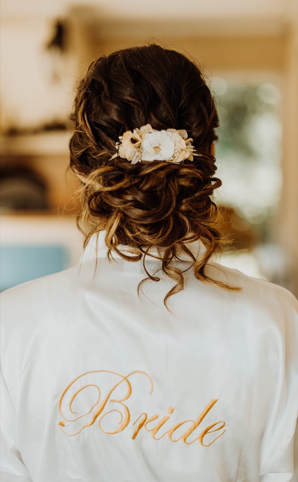 Un chignon flou orné d'un peigne de fleurs blanches, sur une mariée en peignoir brodé Bride